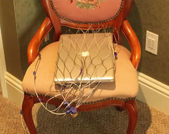 A stainless steel laptop protector with a silver laptop in, is locked to a large wooden chair to prevent theft.